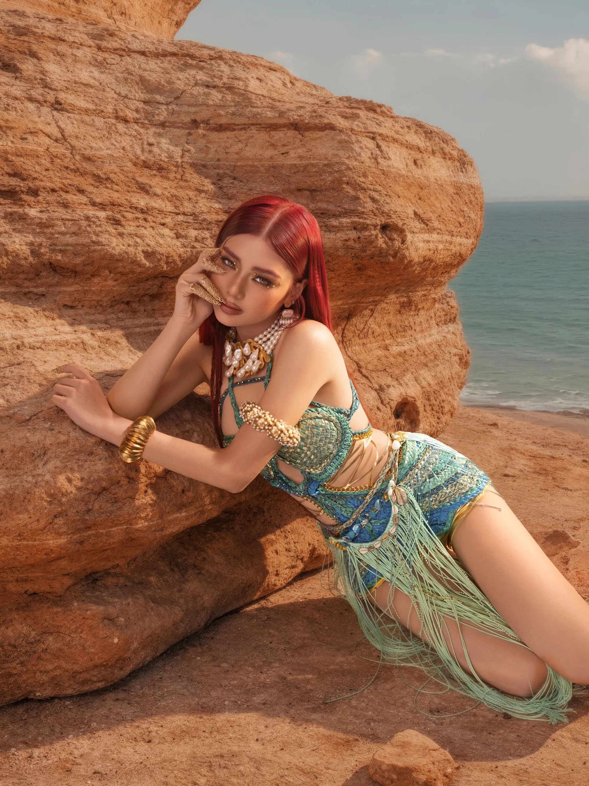 A BINI member with red hair in a beaded teal look, leaning on a sandstone boulder