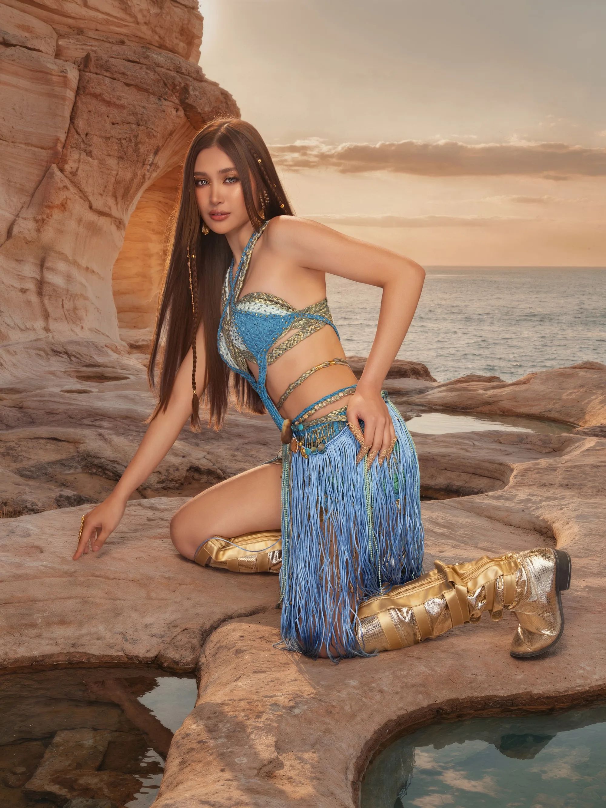 A BINI member with long dark hair in a teal fringe look, kneeling on the rocks at dusk